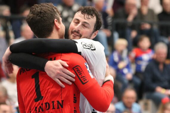 Zwei männliche Sportler umarmen sich auf einem Hallensportplatz, der eine trägt ein rotes Trikot mit der Nummer 1, der andere ein weißes, im Hintergrund ist ein unscharfes Publikum zu sehen.