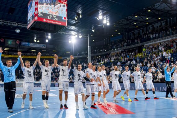 Eine Herren-Handballmannschaft steht in einer Reihe auf einem Hallenplatz, hält sich an den Händen und feiert mit erhobenen Armen, während eine jubelnde Menge zusieht. Auf der Anzeigetafel wird ein Spielstand von 35:24 angezeigt.