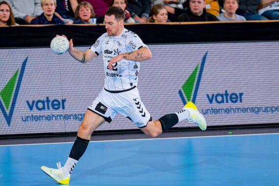 Ein männlicher Handballspieler in einem weiß-schwarzen Trikot springt während eines Spiels nach vorne und hält mit einer Hand einen Ball. Die Zuschauer schauen von der Tribüne aus zu, und im Hintergrund sind Werbetafeln zu sehen.