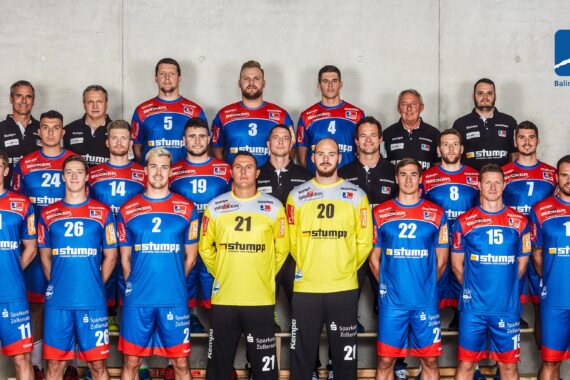 Eine Herren-Handballmannschaft posiert für ein Gruppenfoto in blauen und roten Trikots mit zwei Spielern in gelben Trikots. Sie stehen und knien vor einer grauen Wand mit dem Logo des HBW Balingen-Weilstetten in der rechten oberen Ecke.