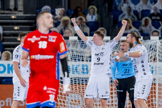 Ein Handballspieler in einem weißen Trikot reißt die Arme hoch, um in der Nähe des Tores zu jubeln, während seine Mannschaftskameraden und ein Torwart sich ihm anschließen. Ein Spieler in einem roten Trikot steht im Vordergrund, während im Hintergrund unscharfe Zuschauer zu sehen sind.