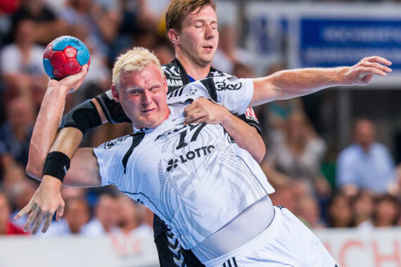 Zwei männliche Handballspieler liefern sich einen intensiven Wettkampf. Der Spieler in Weiß bereitet sich auf den Wurf vor, während der Spieler in Schwarz den Ball abwehrt und über seinen Gegner hinweggreift. Die Szene ist dynamisch, mit einer unscharfen Menschenmenge im Hintergrund.