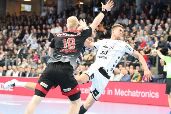 Zwei männliche Handballspieler in Aktion während eines Spiels; einer in einem schwarz-roten Trikot versucht, den anderen in einem weißen Trikot zu blocken oder abzuwehren, während eine große Menschenmenge von der Tribüne aus zusieht.