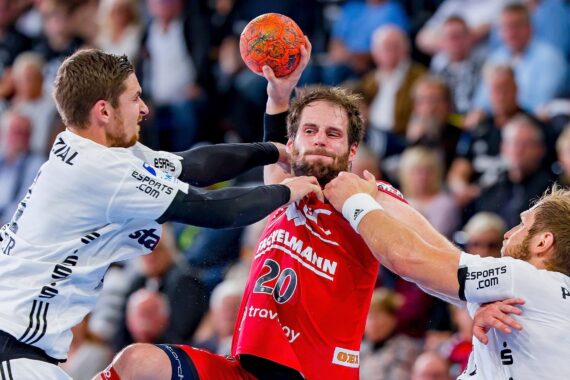 Drei männliche Handballspieler liefern sich einen intensiven Wettkampf; der Spieler in Rot versucht zu schießen, während zwei Spieler in Weiß ihn verteidigen, einer mit einer Hand an seinem Hals, der andere hält seinen Arm fest. Die Menge schaut im Hintergrund zu.