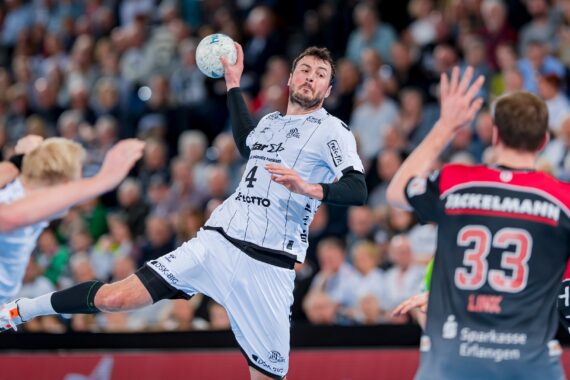 Ein männlicher Handballspieler in einem weißen Trikot springt in die Luft, um den Ball zu werfen, umgeben von gegnerischen Spielern in dunklen Trikots, während eines intensiven Spiels mit einer verschwommenen Menschenmenge im Hintergrund.