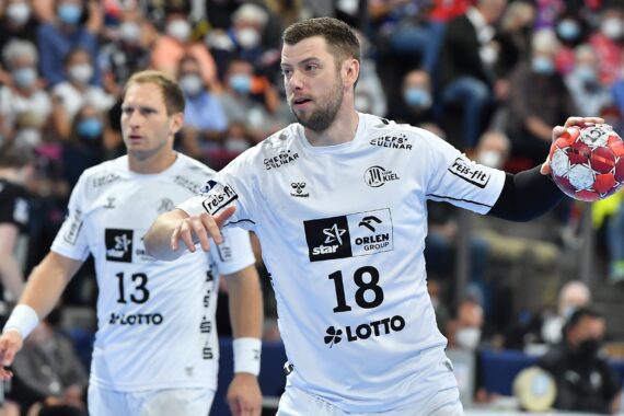 Zwei männliche Handballspieler in weißen Trikots, die Nummer 18 hält einen rot-weißen Ball, die Nummer 13 steht neben ihm. Beide sind auf das Spiel konzentriert, im Hintergrund eine verschwommene Menschenmenge, viele tragen Masken.