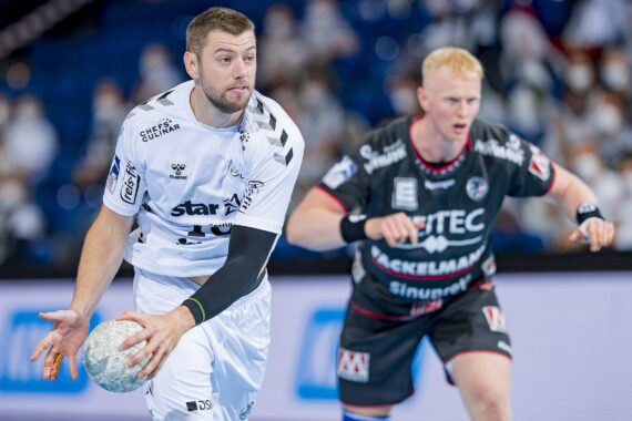 Zwei männliche Handballspieler in Aktion; der Spieler in Weiß hält den Ball, während der Spieler in Schwarz verteidigt. Der Hintergrund zeigt ein unscharfes Publikum und eine Sporthalle.