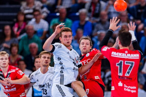 Mehrere männliche Handballspieler in weißen und roten Trikots kämpfen während eines rasanten Spiels um den Ball, während die Zuschauer im Hintergrund zusehen. Ein Spieler in Rot (#17) hebt die Arme, um den Ball abzublocken.