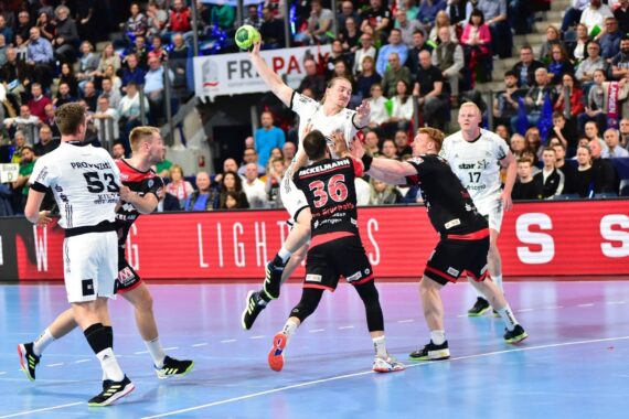Ein Handballspieler in einem weißen Trikot springt, um den Ball in Richtung Tor zu werfen, während mehrere Verteidiger in schwarzen Trikots versuchen, ihn zu blockieren. Eine Zuschauermenge schaut von der Tribüne im Hintergrund zu.
