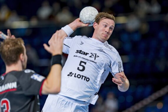 Ein männlicher Handballspieler in einem weißen Trikot mit der Nummer 5 bereitet sich darauf vor, den Ball zu werfen, während ein gegnerischer Spieler in einem dunklen Trikot versucht, ihn während eines Spiels zu blockieren.