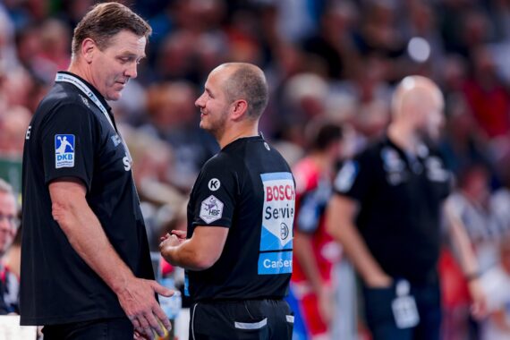 Zwei Schiedsrichter in schwarzen Uniformen unterhalten sich während einer Sportveranstaltung, während im Hintergrund eine unscharfe Menge und andere Offizielle zu sehen sind. Ein Schiedsrichter gestikuliert mit seiner Hand, während der andere aufmerksam zuhört.