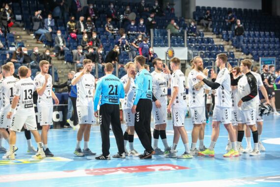 Eine Handballmannschaft in weißen Trikots versammelt sich auf einem Hallenplatz, einige Spieler klatschen sich ab. Die Tribünen hinter ihnen sind mit sitzenden Zuschauern gefüllt.
