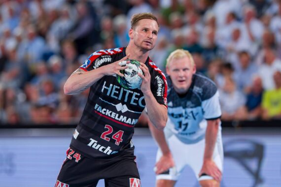 Ein Handballspieler in einem schwarz-roten Trikot mit der Nummer 24 bereitet sich auf den Wurf vor, während ein Spieler in einem weißen Trikot hinter ihm auf dem Spielfeld steht. Im Hintergrund ist eine unscharfe Zuschauermenge zu sehen.