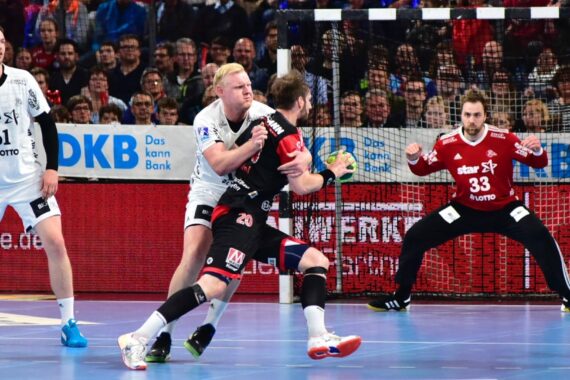 Zwei Handballspieler in Aktion, einer in Weiß verteidigt gegen einen anderen in Schwarz, der gerade auf das Tor schießen will. Der Torwart in Rot ist bereit, während die Zuschauer im Hintergrund aufmerksam zusehen.