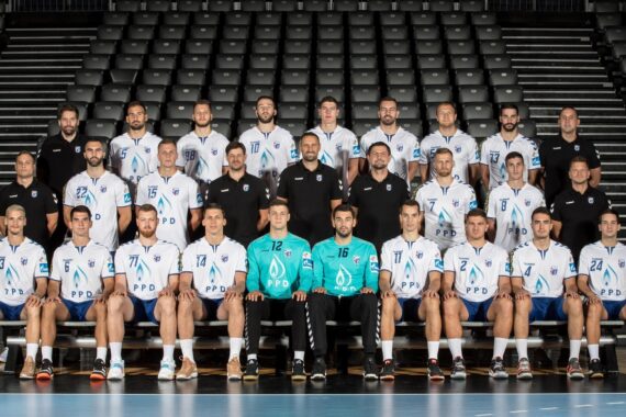 Eine Herrenhandballmannschaft in weißen Trikots stellt sich in einer Halle mit leeren schwarzen Sitzen in drei Reihen sitzend und stehend mit schwarz gekleideten Trainern für ein Gruppenfoto auf.