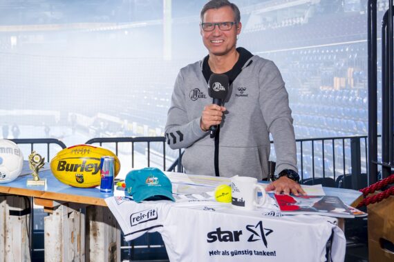 Ein lächelnder Mann mit Brille und grauem Kapuzenpulli steht an einem Tisch mit Sportartikeln, Trophäen, einer Red Bull-Dose und einem weißen Trikot in einer großen, leeren Sportarena.