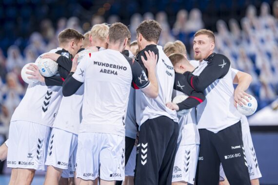 Eine Männerhandballmannschaft in weißen und schwarzen Trikots kauert auf einem Hallenplatz zusammen und hält Handbälle, während im Hintergrund unscharfe Zuschauer zu sehen sind.