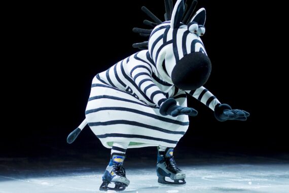 Eine Person in einem verspielten Zebrakostüm läuft mit schwarzen und weißen Streifen, einer großen Schnauze und blauen Schlittschuhen auf dem Eis, wobei die Szene dramatisch ausgeleuchtet wird.