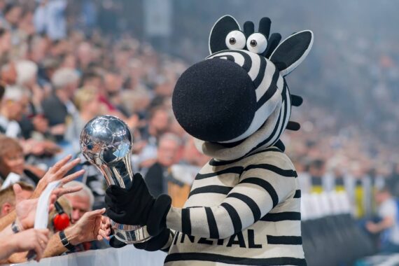 Eine Person in einem Zebra-Maskottchenkostüm hält eine glänzende silberne Trophäe, während sie mit jubelnden Fans in einem überfüllten Stadion interagiert.