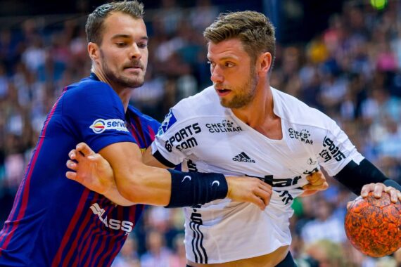 Zwei männliche Handballspieler kämpfen während eines Spiels um den Ball. Der Spieler in Weiß hält den orangefarbenen Ball, während der Spieler in Blau und Rot versucht, ihn zu verteidigen. Im unscharfen Hintergrund sieht eine Menschenmenge zu.