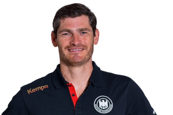 Ein Mann mit kurzen braunen Haaren und heller Gesichtsbehaarung lächelt in die Kamera. Er trägt ein dunkles Poloshirt mit dem deutschen Staatswappen und dem Kempa-Logo auf der Schulter vor einem schlichten weißen Hintergrund.