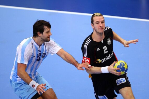 Zwei männliche Handballspieler in Aktion; der eine im schwarzen Trikot versucht, mit dem Ball vorzudringen, während der andere im blau-weißen Trikot versucht, ihn auf einem Hallenplatz zu blockieren.