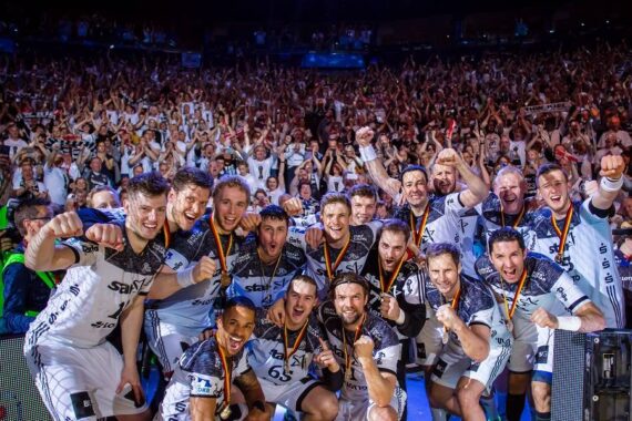 Eine jubelnde Handballmannschaft posiert gemeinsam auf dem Spielfeld, trägt Medaillen und weiße Trikots und feiert ihren Sieg, während eine große, jubelnde Menge die Tribüne hinter ihnen füllt.