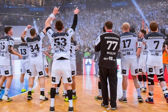 Eine Herren-Handballmannschaft in weißen Trikots feiert auf einem Hallenplatz, mit erhobenen Armen vor einer jubelnden Menge. Konfetti liegt in der Luft, und die Atmosphäre ist lebhaft und festlich.