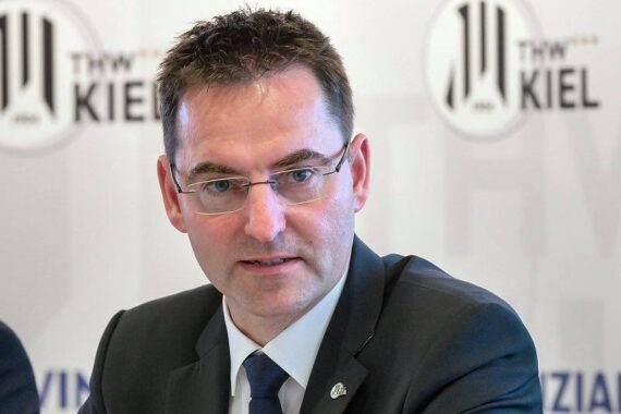 Ein Mann im Anzug und mit Brille spricht bei einer Presseveranstaltung, während im Hintergrund die Logos des THW Kiel zu sehen sind.