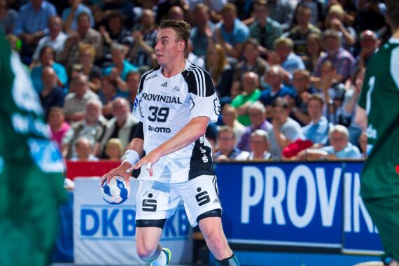 Ein männlicher Handballspieler in einem weiß-schwarzen Trikot, der einen Ball in der Hand hält, bereitet sich auf einen Spielzug während eines Hallenspiels vor. Die Zuschauer füllen die Tribünen im Hintergrund. Die Spieler in Grün sind teilweise im Vordergrund zu sehen.