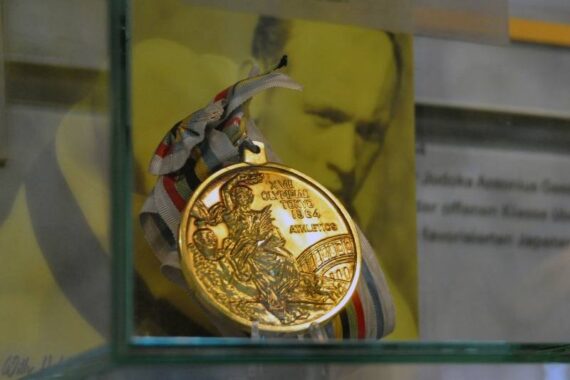 Eine goldene olympische Medaille mit einem Band wird in einer Glasvitrine ausgestellt, wobei ein eingraviertes Muster und die Jahreszahl 1964 sichtbar sind. Im Hintergrund sind ein verschwommenes Porträt und ein Text zu sehen.
