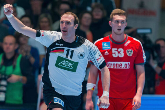 Ein Handballspieler im weiß-schwarzen Deutschland-Trikot hebt jubelnd den Arm, während ein Spieler im roten Österreich-Trikot hinter ihm auf dem Spielfeld steht, mit einer Menschenmenge im Hintergrund.