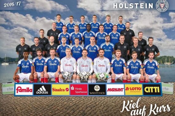 Eine Fußballmannschaft posiert in drei Reihen im Freien, in blau-weißen Trikots, mit Trainern in Schwarz hinter ihnen. Am unteren Bildrand sind Sponsorenlogos zu sehen, und im Hintergrund sind Wasser und Bäume zu sehen.