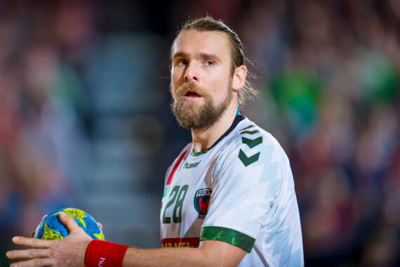 Ein männlicher Handballspieler mit Bart und zurückgebundenen langen Haaren hält einen blau-gelben Handball. Er trägt ein weißes Trikot mit der Nummer 28 und scheint während eines Spiels konzentriert zu sein, mit unscharfen Zuschauern im Hintergrund.