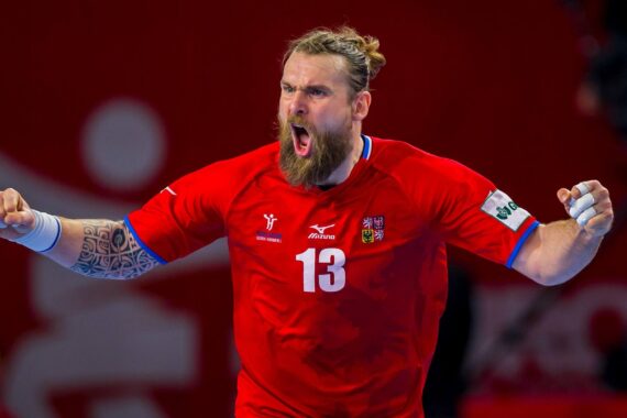 Ein männlicher Handballspieler mit Bart und tätowiertem Arm, der ein rotes Trikot mit der Nummer 13 trägt, jubelt leidenschaftlich mit geballten Fäusten vor einem roten Hintergrund.