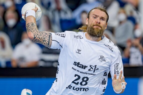 Ein männlicher Handballspieler mit Bart und tätowiertem Arm, der ein weißes Trikot mit der Nummer 28 trägt, ist mitten in der Aktion, um den Ball während eines Spiels zu werfen, wobei die Zuschauer im Hintergrund verschwommen sind.