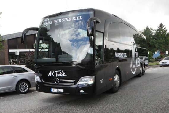 Ein schwarzer Mercedes-Benz Reisebus mit der Aufschrift "Wir sind Kiel" steht auf einer Straße neben einem silbernen Auto, im Hintergrund sind Bäume und Gebäude zu sehen.