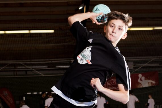 Ein Junge in einem schwarzen Sportdress bereitet sich darauf vor, während eines Hallenhandballspiels einen Ball zu werfen, während im Hintergrund andere Spieler und ein Netz zu sehen sind.