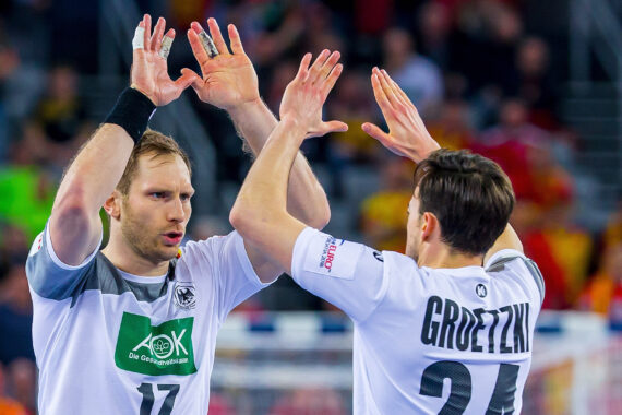 Zwei männliche Handballspieler in weißen Trikots, einer mit 17 und einer mit 24 mit der Aufschrift GROETZKI, klatschen während eines Spiels in die Hände. Im Hintergrund sind unscharfe Zuschauer und eine hell erleuchtete Halle zu sehen.