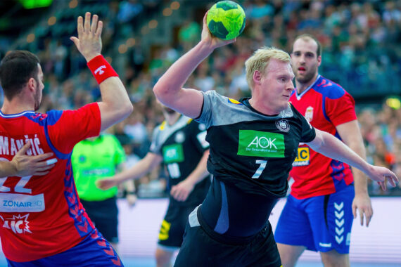 Ein männlicher Handballspieler in einem schwarzen Trikot bereitet sich darauf vor, den Ball zu werfen, während seine Gegner in roten Trikots versuchen, ihn während eines intensiven Hallenspiels zu blockieren. Die Zuschauer schauen von den Tribünen im Hintergrund zu.
