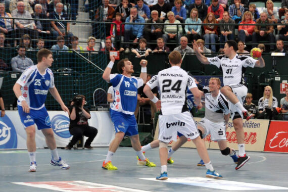 Eine Gruppe männlicher Handballspieler in blau-weißen Trikots ist auf einem Hallenplatz in Aktion, wobei ein Spieler den Ball in der Luft hält. Die Zuschauer schauen von der Tribüne im Hintergrund zu.
