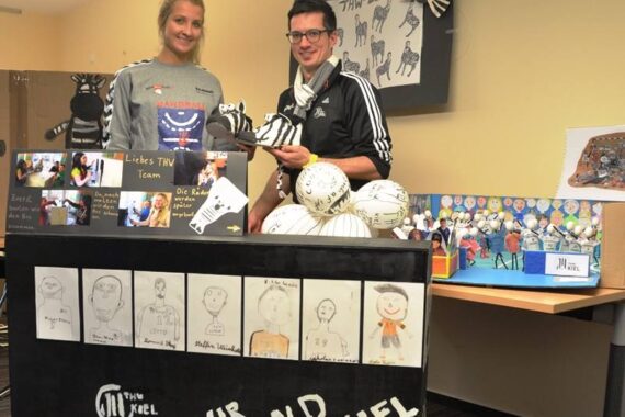 Zwei Personen stehen hinter einem schwarzen Display mit Kinderzeichnungen und dem Text Wir sind Kiel. Auf dem Tisch liegen auch verzierte Fußbälle, kleine Basteleien und Fotos, während im Hintergrund weitere Kunstwerke und Dekorationen zu sehen sind.