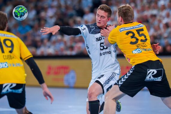 Drei männliche Handballspieler kämpfen während eines Spiels um den Ball. Ein Spieler in Weiß spielt einen Pass oder schießt, während zwei Spieler in Gelb ihn verteidigen. Die Zuschauer sind im Hintergrund verschwommen.