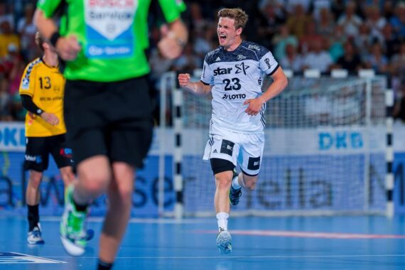Ein männlicher Handballspieler in einem weißen Trikot jubelt auf dem Platz, ballt die Faust und schreit, während im Hintergrund andere Spieler und ein unscharfer Schiedsrichter in Grün zu sehen sind.