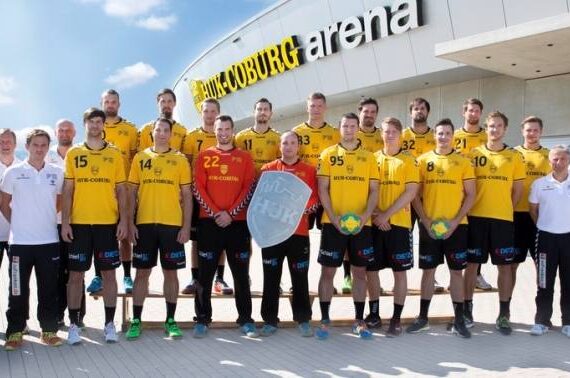 Eine Männer-Handballmannschaft in gelben Trikots und schwarzen Shorts posiert mit Trainern und Mitarbeitern vor einem modernen Gebäude mit der Bezeichnung Arena unter blauem Himmel. Einige Spieler halten Handbälle und ein Schild.