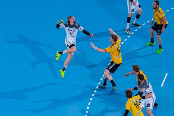 Ein Handballspieler in einem weißen Trikot springt, um den Ball in Richtung Tor zu schießen, während Spieler in gelben Trikots auf einem blauen Hallenplatz verteidigen.