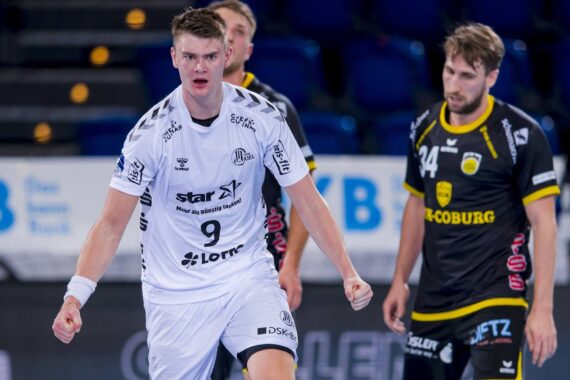 Ein männlicher Handballspieler in einem weißen Trikot jubelt energisch auf dem Spielfeld, während zwei Spieler in schwarzen Trikots während eines Spiels im Hintergrund zusehen.
