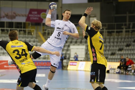 Ein Handballspieler in einem weißen Trikot springt, um den Ball zu schießen, während zwei Verteidiger in gelben und schwarzen Trikots versuchen, ihn auf einem Hallenplatz zu blockieren. Die Tribünen im Hintergrund sind größtenteils leer.