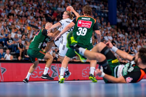 Ein Handballspieler in einem weißen Trikot springt, um den Ball zu werfen, während ein Verteidiger in einem grünen Trikot versucht, ihn zu blockieren. Zwei weitere grün gekleidete Spieler befinden sich auf dem Boden. Die Aktion findet vor einer großen, jubelnden Menge statt.
