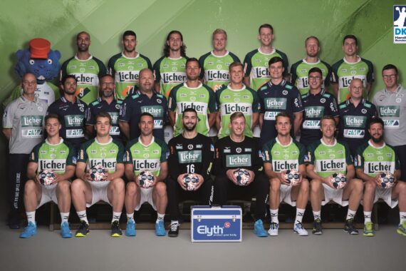 Eine Herren-Handballmannschaft und Mitarbeiter posieren für ein Gruppenfoto in grün-schwarzen Uniformen, halten Handbälle und lächeln. Ein blaues Maskottchen steht links und eine Kühlbox mit der Aufschrift Elyth steht vorne in der Mitte. Das Logo der DKB Handball-Bundesliga ist oben rechts zu sehen.